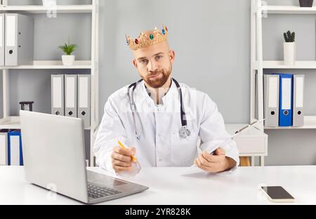 Lustiger junger Arzt in weißer medizinischer Uniform am Arbeitsplatz in Klinik mit Krone. Stockfoto
