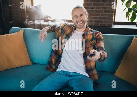 Entspannter junger Mann in kariertem Hemd, der Freizeit auf dem Sofa in stilvollem Wohnzimmer mit hellem Tageslicht genießt Stockfoto