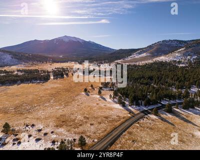 Hohe Luftaufnahme einer Bergstraße, die sich durch eine verschneite Wiese und einen Kiefernwald zum Humphreys Peak hin schlängelt, unter einer hellen Sonne in der Nähe von Flagstaff Stockfoto