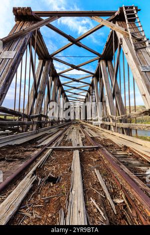 Die Brücke ist alt und verrostet, mit einer Holzkonstruktion und einer Metallschiene. Der Himmel ist blau und klar und die Brücke ist von Bäumen umgeben Stockfoto