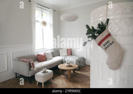 Weihnachtsstrumpf, grüne Tannenzweige hängen am alten Kachelofen. Winterlich dekoriertes skandinavisches Wohnzimmer. Sofa, rote Kissen, Kaffee Stockfoto