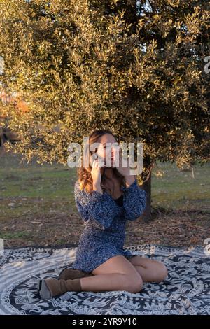 Eine hübsche junge Frau in einem blauen Kleid lacht, indem sie ihre Hand über den Mund legt, während sie auf ihrem Handy sitzt und in einem Mandala mitten in Natu sitzt Stockfoto