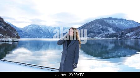 Eine fröhliche Frau in Mantel und Fäustlingen lächelt, während sie den beschaulichen schneebedeckten Fjord und die Berge in Gaupne, Norwegen, bewundert Stockfoto