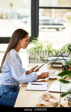 Eine junge Frau mit langen brünetten Haaren sitzt an einem Holztisch, schlürft Kaffee und benutzt ihr Telefon. Stockfoto