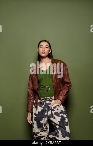 Selbstbewusste junge Frau in brauner Lederjacke und gemusterter Hose lehnt sich an eine grüne Wand. Stockfoto