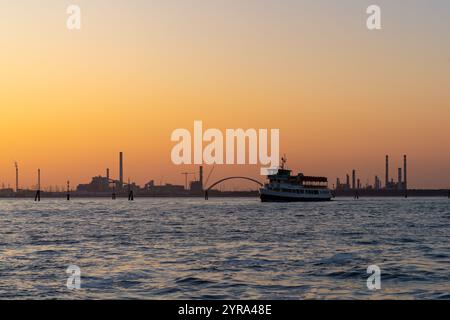 Eine Pasengerfähre im Giudecca-Kanal mit der Ponte Bossi- oder Bossi-Brücke dahinter in Venedig, Italien. Auf der linken Seite befindet sich ein Kraftwerk mit einer Ölraffinerie bei Stockfoto