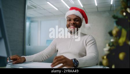 Marketing Manager oder Finanzspezialist arbeiten am Computer in modernen Büros. Junger erfolgreicher afroamerikanischer Geschäftsmann in Santa hat sitzt am Arbeitsplatz, lächelt und schaut in die Kamera. E-Commerce. Stockfoto