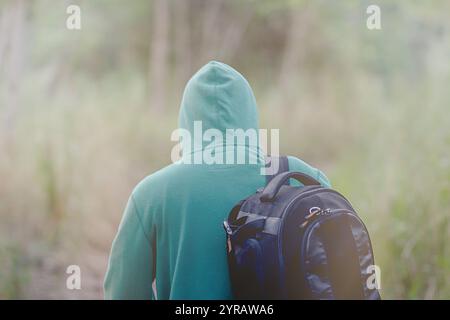 Der Rücken des jungen Fotografen trägt grünen Hoodie, der mit schwarzer Kameratasche auf dem Weg durch den Wald läuft, mit verschwommenem Hintergrund der grünen Natur im Winter Stockfoto