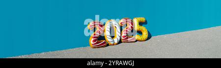 Einige Folienballons in verschiedenen Farben bilden die Zahl 2025, wie das neue Jahr, auf dem Asphalt, gestützt auf eine blaue Mauer im Freien, in einer Panoramaform Stockfoto