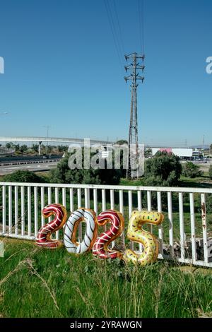 Vier zahlenförmige Ballons in verschiedenen Farben bilden die Zahl 2025 als neues Jahr, die auf einem weißen Geländer in einem Industriegebiet, auf einer sonnigen Fläche, gestützt auf ein weißes Geländer Stockfoto