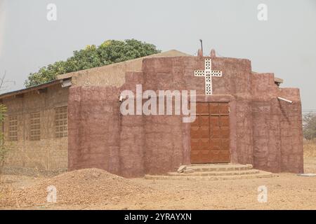 Rustikale Clay Village Church mit Kreuz und Holztür im ländlichen Togo, umgeben von trockener Landschaft Stockfoto