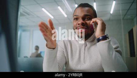 Begeisterte junge Geschäftsmann freut sich und feiert erfolgreiche Geschäftsabschlüsse mit Kunden, nutzt Computer, spricht per Mobiltelefon. Afroamerikaner Marketingspezialist arbeitet in modernen Büros. E-Commerce Stockfoto