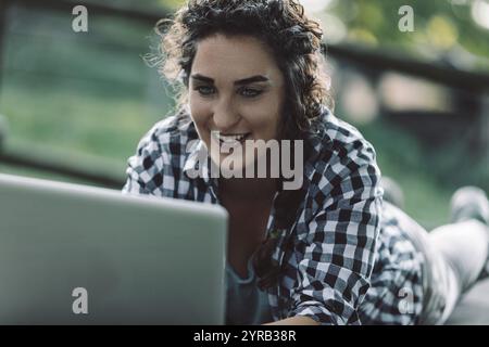 Eine junge Frau mit lockigen Haaren, die ein kariertes Hemd trägt, liegt draußen auf ihrem Bauch, lächelt, während sie an ihrem Laptop arbeitet und vielleicht das frische genießt Stockfoto