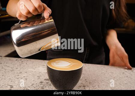 Barista gießt aufgeschäumte Milch aus dem Pitcher in eine Tasse aromatischen Kaffee im Café, Nahaufnahme. Stockfoto