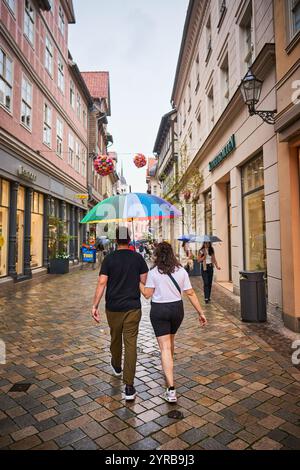 Ein Paar geht Hand in Hand unter einem bunten Regenschirm entlang einer kopfsteingepflasterten Straße in Schottland, mit Geschäften entlang der Straße und einem regnerischen Ambiente. Stockfoto