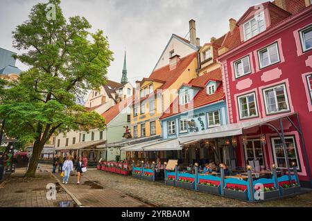 Eine lebhafte Straßenszene in Riga, Lettland, mit farbenfrohen Gebäuden und Restaurants im Freien, umgeben von üppigem Grün und einem bewölkten Himmel. Stockfoto