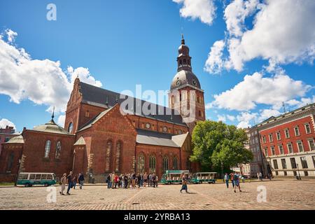 Eine lebendige Szene in Riga, Lettland, mit einer historischen Backsteinkirche mit einem hohen Uhrenturm, umgeben von einem kopfsteingepflasterten Platz mit Besuchern und Stockfoto