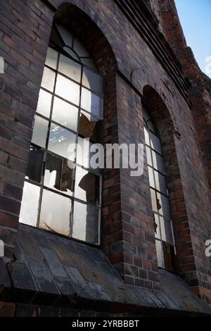 Eine Nahaufnahme aus zersplittertem Glas in den Fenstern eines verlassenen Industriegebäudes aus Ziegelstein vor einer düsteren städtischen Kulisse. Stockfoto