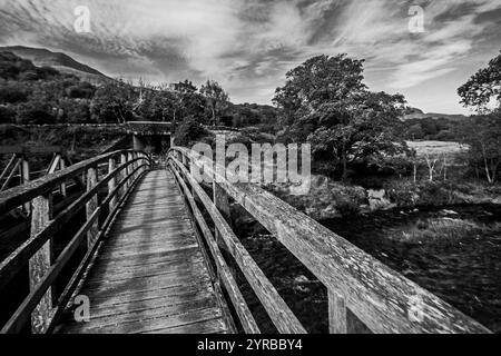 Dramatische Schwarzweiß-Aussicht entlang einer verwitterten hölzernen Wanderbrücke über den Fluss Glaslyn im Eryri-Nationalpark in Wales Stockfoto