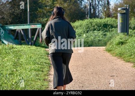 Eine junge Frau mit Kopfhörern spaziert durch einen üppigen Park und genießt Musik und frische Luft. Von hinten fotografiert, zeigt diese ruhige Szene Outdo Stockfoto