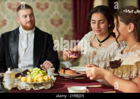 Seitenansicht Porträt eines jungen Herrn im Samtfrack, der elegante Damen am Esstisch in klassischer Inneneinrichtung ansieht, beleuchtet durch warme Beleuchtung Stockfoto