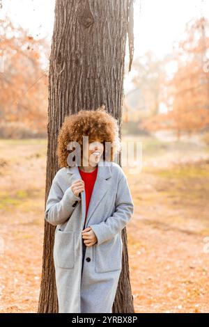 Eine junge Frau mit lockigen Haaren lehnt sich an einen Baum, trägt einen warmen Pullover, lächelt fröhlich im goldenen Herbstlaub und umhergefallenen Blättern Stockfoto