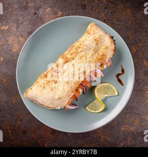 Geschlossene Calzone-Pizza mit Räucherlachs, Frischkäse, roten Zwiebeln und Zitronenscheiben, serviert auf einem eleganten blauen Teller Stockfoto