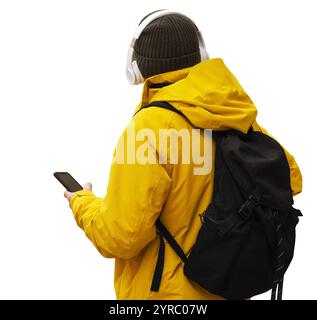 Isolierte Rückansicht einer Person mit gelber Winterjacke und Hut, stehend mit Mobiltelefon, kabellosen Kopfhörern und Rucksack Stockfoto