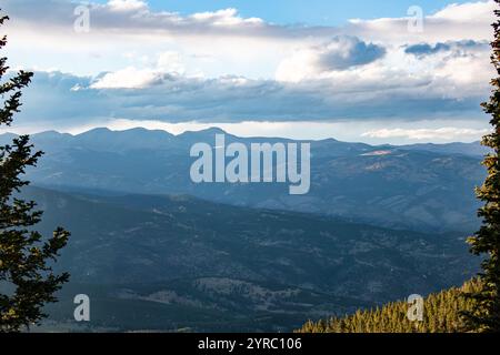 Ein Panoramablick auf entfernte Bergketten, eingerahmt von immergrünen Bäumen, mit einem bewölkten Abendhimmel, der Schatten über das Gelände wirft. Stockfoto