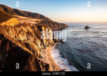 Goldene Küstenklippen in Big Sur, Kalifornien, mit Wellen, die an einem Sandstrand und einem Horizont bei Sonnenuntergang plätschern. Stockfoto