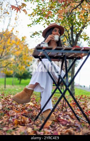 Bei einem warmen Getränk im Freien sitzt eine junge lateinfrau an einem kleinen Tisch inmitten gefallener Herbstblätter und genießt die ruhige Atmosphäre eines Parks Stockfoto