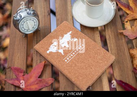 Das Notizbuch mit Weltkarte und Adventures-Inschrift liegt auf einem Holztisch mit herabfallenden Herbstblättern, neben einer Tasse Kaffee und einem Wecker, sugg Stockfoto
