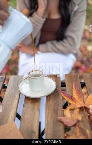 Die Person gießt Kaffee aus einer Mokakanne in eine weiße Tasse auf einem Holztisch mit Herbstlaub in einem Park, wodurch eine gemütliche und saisonale Atmosphäre entsteht Stockfoto