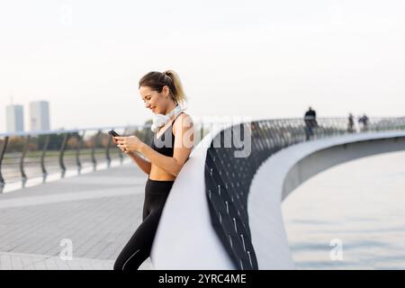 Eine Joggerin hält an ihrem Telefon am Fluss an, trägt Trainingsausrüstung und Kopfhörer und genießt die warme Sonne und die ruhige Atmosphäre Stockfoto