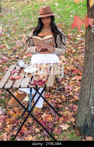 Die junge lateinfrau mit Hut sitzt an einem Holztisch in einem Park, der mit Herbstlaub bedeckt ist, genießt eine Tasse Kaffee und liest ein Buch darüber Stockfoto