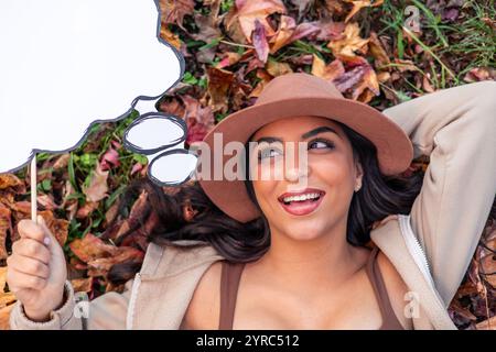 Lächelnde junge Frau mit braunem Hut liegt auf einem Bett aus Herbstlaub, hält eine leere Sprechblase und blickt träumerisch weg Stockfoto