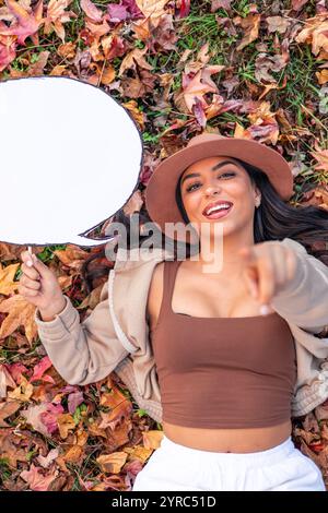 Die junge lateinfrau mit braunem Hut und braunem Oberteil liegt auf Herbstlaub und hält eine leere Sprechblase, während sie mit dem Finger zeigt Stockfoto