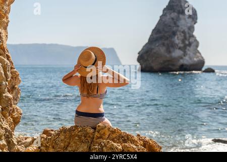 Woman Beach Rock – Eine Frau mit Bikini und Hut sitzt auf einem Felsen und bewundert den Blick auf einen Strand und eine große Felsformation in der Ferne. Stockfoto