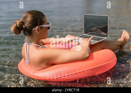 Eine Frau sitzt auf einem pinkfarbenen Schlauchboot im Wasser und benutzt einen Laptop. Das Konzept der Entspannung und Freizeit, während die Frau ihre Zeit am genießt Stockfoto