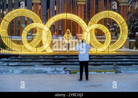 Die Person fotografiert einen anderen, der während des Winters in einer schneebedeckten städtischen Umgebung in der Dämmerung vor beleuchteten Ringen steht. Calgary Olympic Plaza Stockfoto