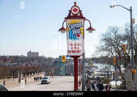 Straßenschild für Clifton Hill, „The Street of Fun by the Falls“, mit Niagarafällen und Fußgängern im Hintergrund an einem sonnigen Tag. Stockfoto