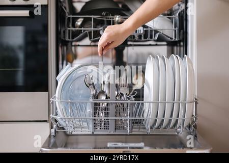 Vorderansicht von der weiblichen Hand, um sauberes Besteck aus einem beladenen Geschirrspüler zu entfernen. Hintergrund der modernen Küche Stockfoto