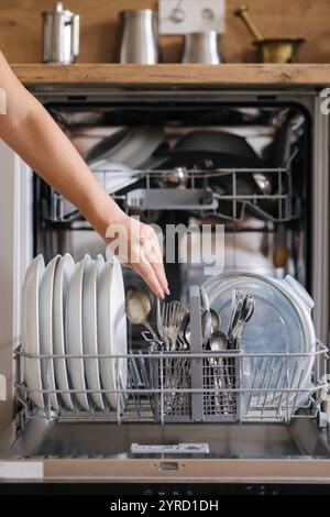 Vorderansicht von der weiblichen Hand, um sauberes Besteck aus einem beladenen Geschirrspüler zu entfernen. Hintergrund der modernen Küche Stockfoto