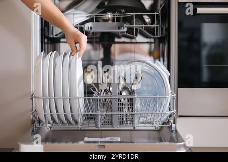 Vorderansicht von weiblicher Hand, Entfernen der sauberen Platte aus einem beladenen Geschirrspüler. Hintergrund der modernen Küche Stockfoto