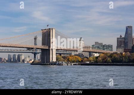 Die Wahrzeichen-Brücke steht vor einem klaren Himmel und verbindet Manhattan mit Brooklyn. Bäume mit Herbstlaub säumen den Wasserrand Stockfoto