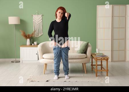 Verärgerte junge übergewichtige Frau in engen Jeans, die zu Hause den Daumen nach unten zeigt Stockfoto