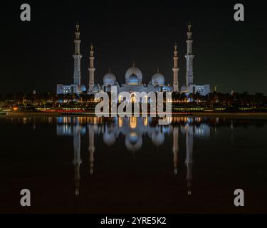 Die große Moschee von Scheich Zayed, die bei Nacht beleuchtet wird, ihre atemberaubende Architektur spiegelt sich in ruhigem Wasser wider und repräsentiert islamische kulturelle Pracht Stockfoto