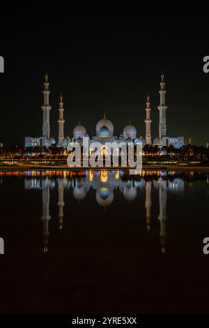 Die große Moschee von Scheich Zayed, die bei Nacht beleuchtet wird, ihre atemberaubende Architektur spiegelt sich in ruhigem Wasser wider und repräsentiert islamische kulturelle Pracht Stockfoto