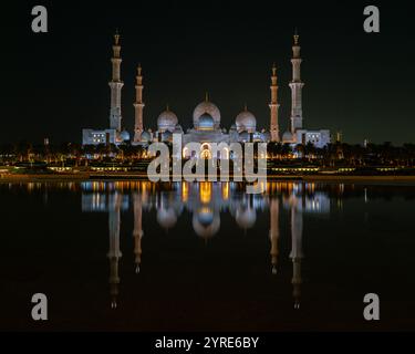 Die große Moschee von Scheich Zayed, die bei Nacht beleuchtet wird, ihre atemberaubende Architektur spiegelt sich in ruhigem Wasser wider und repräsentiert islamische kulturelle Pracht Stockfoto