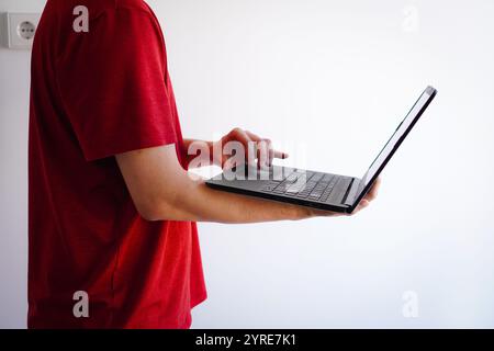 Junger männlicher Investor in rotem T-Shirt, der in einem Zimmer mit weißen Wänden mit Laptop steht und im Internet surft Stockfoto